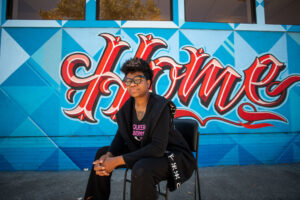 HAC outreach advocate sits on a folding chair in front of the West Oakland HAC mural. The mural text reads “Home”.